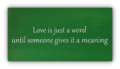 A green wooden plaque with white vinyl letters displaying the phrase 'Love is just a word until someone gives it a meaning'.
